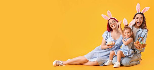 Feliz pareja lésbica con hija pequeña y huevos de Pascua sobre fondo amarillo con espacio para texto - Foto, Imagen