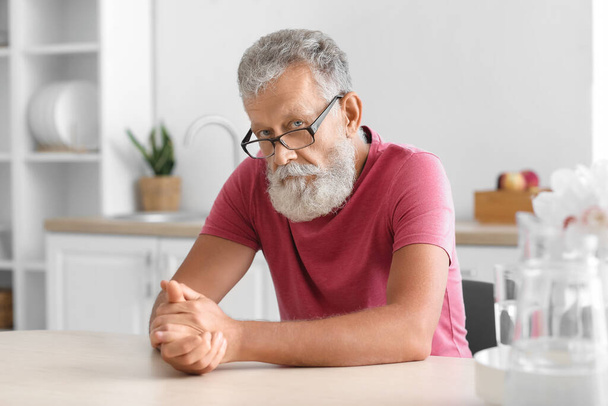 Senior bearded man with eyeglasses sitting at table in kitchen - Fotografie, Obrázek
