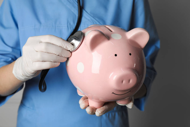 Doctor holding pale pink ceramic piggy bank with stethoscope against white wall, closeup. Medical insurance - Photo, image