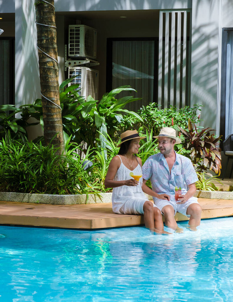 Şapkalı erkekler ve kadınlar tatil sırasında havuz kenarında kokteyl içerler. Taylandlı kadınlar ve beyaz erkekler yüzme havuzunda dinleniyorlar. - Fotoğraf, Görsel