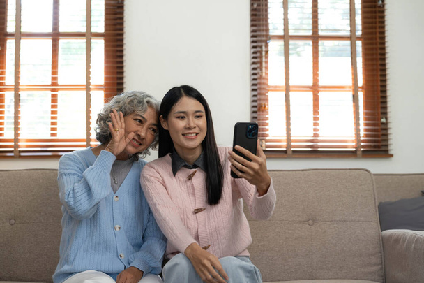 Smiling older mother and adult daughter using phone together, sitting on cozy sofa at home, happy young woman and mature mum looking at smartphone screen, watching video, having fun, two generations.. - Photo, Image