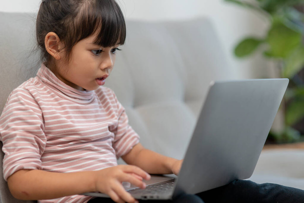 Aasialainen pieni koulutyttö käyttää kannettavaa tietokonetta sohvalla yksin kotona. Lapsen oppiminen käsittelyssä verkossa sosiaalisen median sisältöä, pelata opetus oppitunteja peli chattailuun ystävien kanssa. Lapset tech riippuvuus - Valokuva, kuva