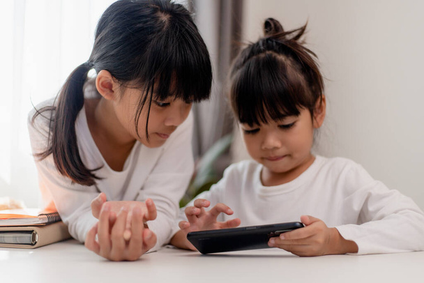 Lapsia ja vempaimia. Kaksi pientä tyttöä sisarukset sisarukset katsoa puhelimeen ja hymyillä. Heillä on älypuhelin katsella videoita, oppia, pelata pelejä, puhua verkossa. Internet lapsille. Vanhempainvalvonta - Valokuva, kuva