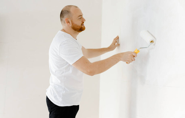 Giovane uomo dipinge il muro con un rullo con vernice bianca. il concetto di ristrutturazione della casa, ristrutturazione nell'appartamento - Foto, immagini
