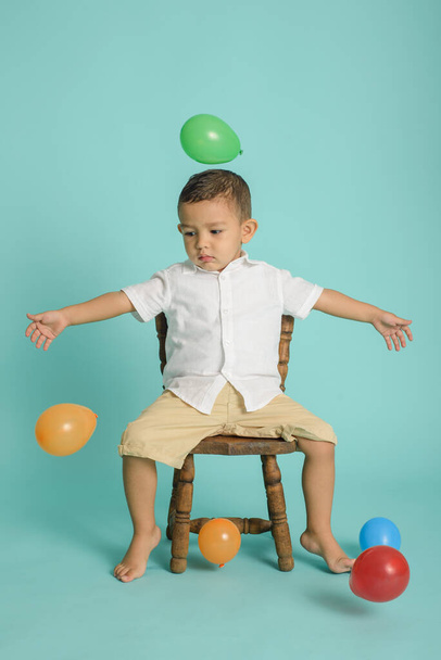Retrato de menino vestido de branco brincando com balões sentados em cadeira de madeira. Fundo azul. - Foto, Imagem