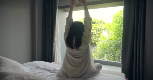 Οπίσθια όψη μιας γυναίκας κάνει τέντωμα μετά το ξύπνημα το πρωί, κοιτάζοντας το πράσινο θέα της φύσης έξω από το παράθυρο του υπνοδωματίου - Πλάνα, βίντεο