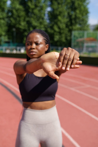 Young woman in black top stands on red track warming up before intensive workout. African American female sports beginner stretches hands against green trees growing in city park on sunny summer day - Φωτογραφία, εικόνα