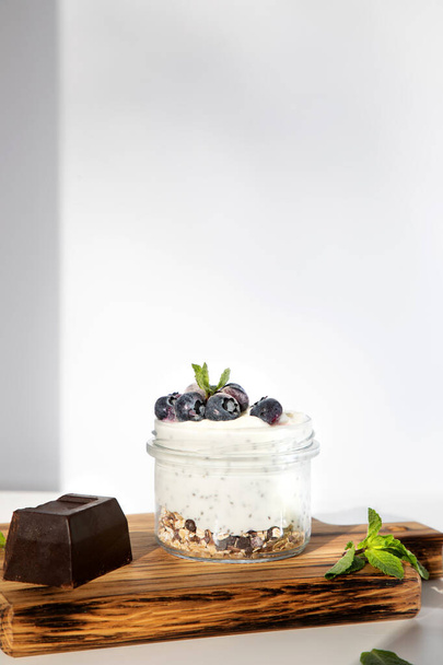 グラノーラ、チョコレート、ブルーベリーとミントの葉と白い背景に木製のボード上の蜂蜜とガラス瓶の中にヨーグルト。正面図 - 写真・画像