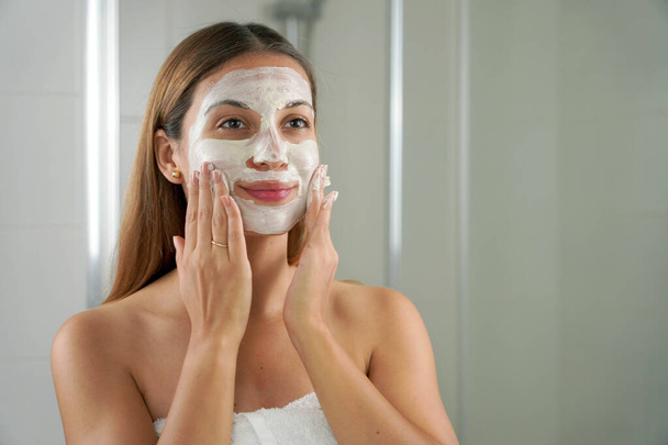 Портрет красивой женщины с глиняной маской на лице, смотрящей в зеркало в ванной комнате - Фото, изображение