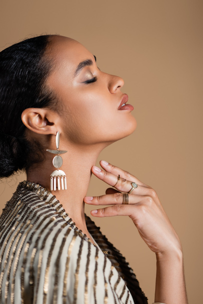 side view of young african american woman in luxurious golden jewelry posing isolated on beige - Photo, Image