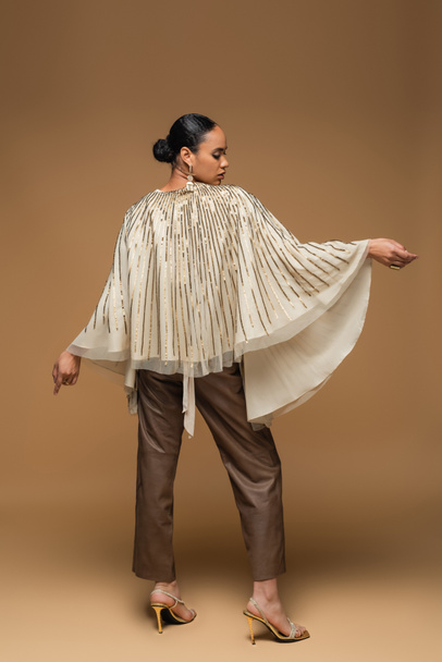 longitud completa de la elegante mujer afroamericana en chal de oro y pantalones de cuero de pie en beige - Foto, imagen