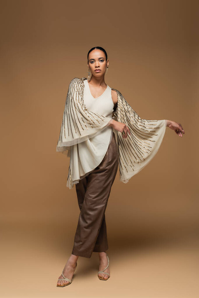 full length of stylish african american woman in golden shawl and leather pants posing on beige - Zdjęcie, obraz