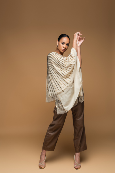 full length of stylish african american model in golden jewelry and shawl posing on beige  - Foto, Imagen