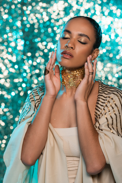 brunette african american woman in shawl and gold on neck posing with closed eyes on sparkling blue background  - 写真・画像