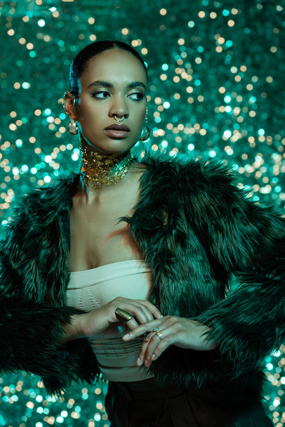 brunette african american woman in faux fur jacket and gold on neck posing on sparkling turquoise background  - Fotografie, Obrázek