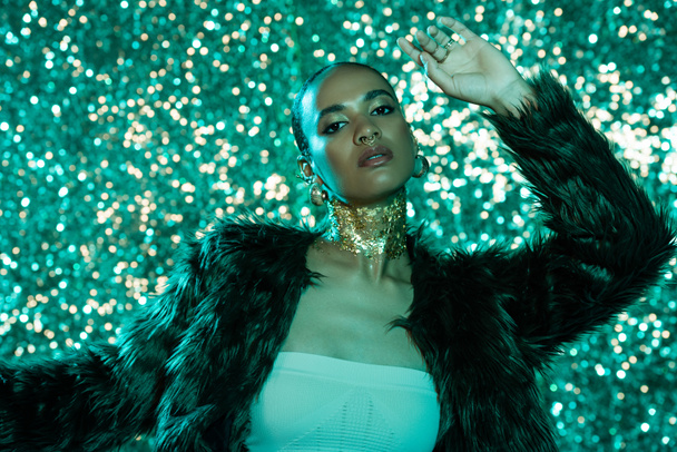 pretty african american woman in faux fur jacket and gold on neck posing on shiny blue background  - Valokuva, kuva