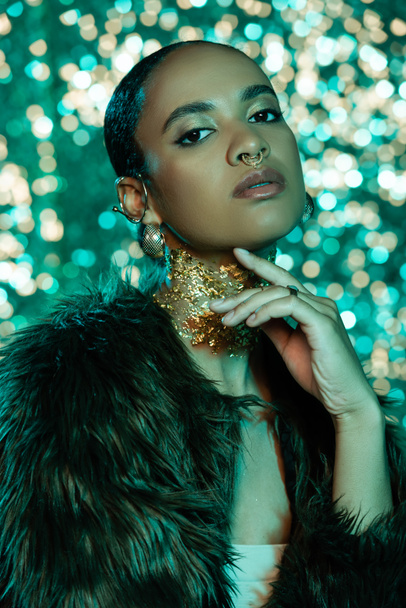 Portrait of fashionable african american woman in faux fur jacket with golden foil on neck posing on sparkling background  - Valokuva, kuva