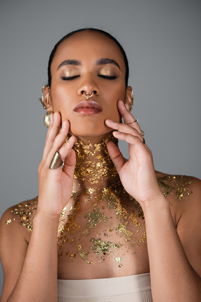 Όμορφη Αφροαμερικανή γυναίκα με χρυσό μακιγιάζ και αλουμινόχαρτο στο στήθος που αγγίζει μάγουλα απομονωμένα σε γκρι  - Φωτογραφία, εικόνα