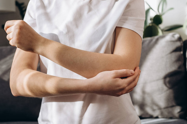 Girl suffering from pain in hand at home. Health care concept. Close up of unhealthy young female sitting on couch having pain in elbow and using hand for self massage to relieve aches and pains - Foto, Bild