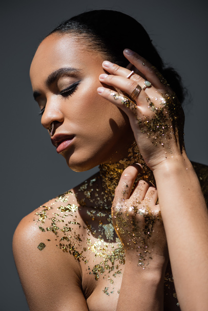 Young african american woman with golden foil on body touching face while posing in light isolated on grey  - Photo, Image