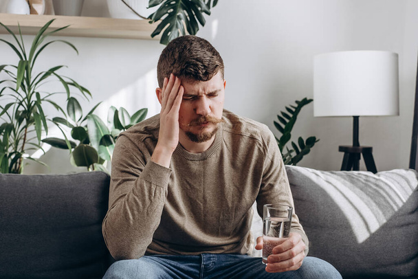 Upset young caucasian man suffering from strong headache or migraine sitting on sofa at home with glass of water, guy feeling intoxication and pain touching aching head. Morning after hangover concept - Foto, Bild