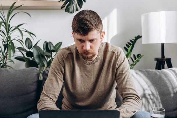 Close up of focused young man sitting on couch with laptop, working remotely online from home. Concentrated millennial male studying distantly alone in living room at home. Freelance concept - Foto, Imagen