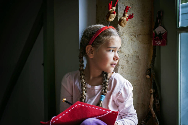 Мечтательная девушка в забавной шляпе пишет Санте письмо и ждет подарков в канун Рождества. Зимние каникулы в разных местах, традиции Ноэля - Фото, изображение
