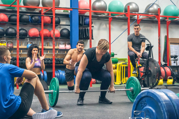 Прикута жінка готова підняти вагу, в той час як її товариші по спортзалу підбадьорюють її
 - Фото, зображення