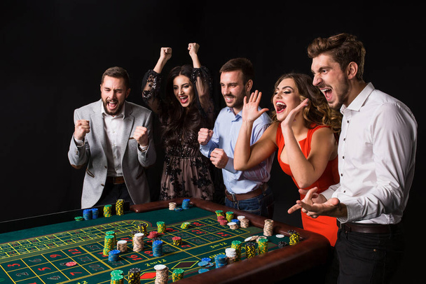 Group of young people behind roulette table on black background. Young people made bets in the game and wait for the result. Bright emotions - Photo, Image
