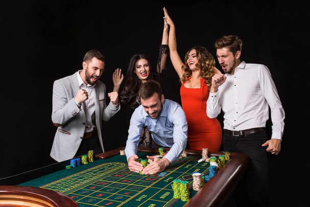 Gruppe junger Leute hinter Roulettetisch auf schwarzem Hintergrund. Gewinner Spieler. Helle Emotionen - Foto, Bild