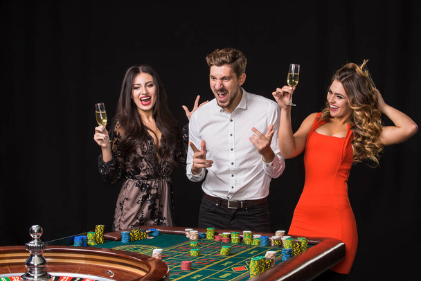 Dos mujeres jóvenes y un hombre detrás de la mesa de ruleta sobre fondo negro. Emociones jugadores - Foto, Imagen