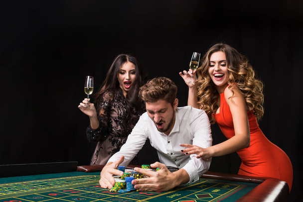 Dos mujeres jóvenes y un hombre detrás de la mesa de ruleta sobre fondo negro. Emociones jugadores - Foto, imagen