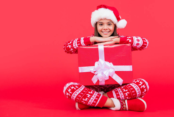 new year. positive santa teen girl with new year present sit with copy space. teen girl hold new year santa present isolated on red background. - Valokuva, kuva