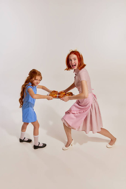 Funny image of beautiful woman, mother and little cute girl, daughter fighting for freshly baked buns over grey background. Concept of beauty, retro style, fashion, 60s, 70s, family. Copy space for ad - Photo, Image