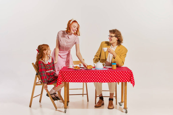 Portrait of beautiful woman, man and little girl sitting at the table and having breakfast isolated over grey background. Concept of beauty, retro style, fashion, 60s, 70s, family. Copy space for ad - Foto, Imagen