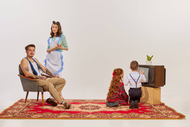 Portrait of young family watching TV isolated over grey background. Kids watching cartoons. Concept of beauty, retro style, fashion, elegance, 60s, 70s, family. Copy space for ad - Фото, изображение