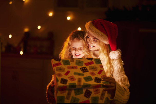 Front view of happy family sitting in decorated room, celebrating new year and christmas. Blonde woman wearing christmas hat opening present with daughter. Concept of winter holiday. - Photo, Image
