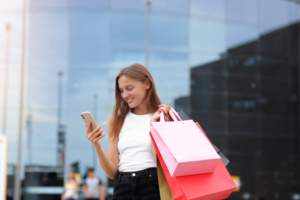 Zwarte vrijdag. Glimlachende jonge vrouw met boodschappentassen gebruikt smartphone op shopping mall achtergrond tijdens het winkelproces, concept van consumentisme, verkoop, rijk leven.Commercieel aanbod voor reclame. - Foto, afbeelding
