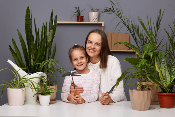 Indoor shot of smiling woman with dark hair posing with cute little daughter in room with greenery, sitting at table, taking care of flowers, looking at camera. - Фото, изображение