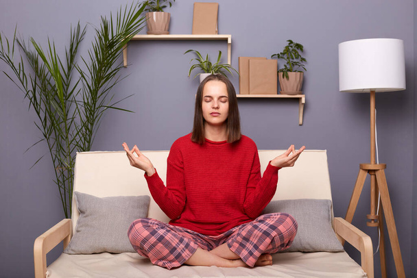 Portrait of calm young adult brown-haired woman wearing red sweater and checkered pants sitting on cough in lotus pose in living room at home, practicing yoga, relaxing. - Foto, afbeelding