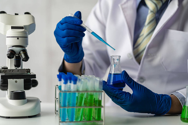 Egy tudós vagy egy orvos laboratóriumi köpenyben tart egy kémcsövet reagens mikroszkóppal és egy csepp színes folyadékkal egy biokémiai laboratóriumi kutatást végző üvegkészüléken.. - Fotó, kép
