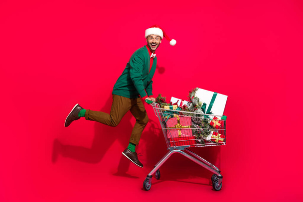 Full length photo of funny excited guy dressed xmas green cardigan buying noel gifts shoppers tray isolated red color background. - Фото, зображення