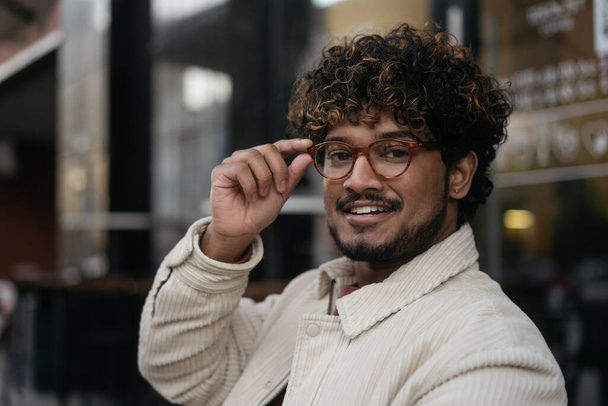 Pozitív göndör hajú ázsiai divat modell pózol képeket az utcán. Jóképű, mosolygós indiai férfi elegáns szemüvegben, aki a szabadban nézi a kamerát. Jövőkép  - Fotó, kép