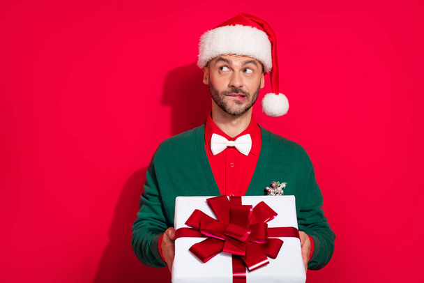 Photo of doubtful unsure guy dressed xmas green cardigan bowtie looking empty space holding noel gift isolated red color background. - Fotó, kép