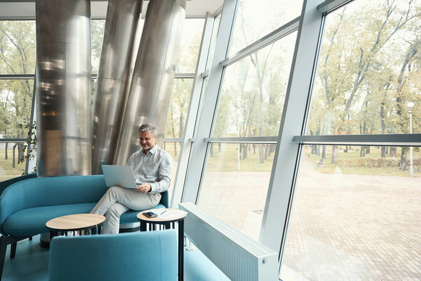 Mann kommuniziert angenehm online mit Kollegen, er sitzt an einem riesigen Sichtfenster in einem Erholungsgebiet - Foto, Bild