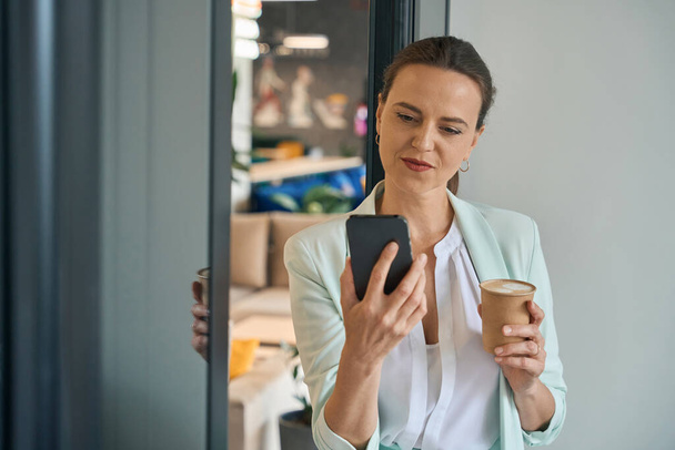 Cheerful elegant lady is standing in office and holding cup of drink in one hand and cell phone in other - Фото, изображение