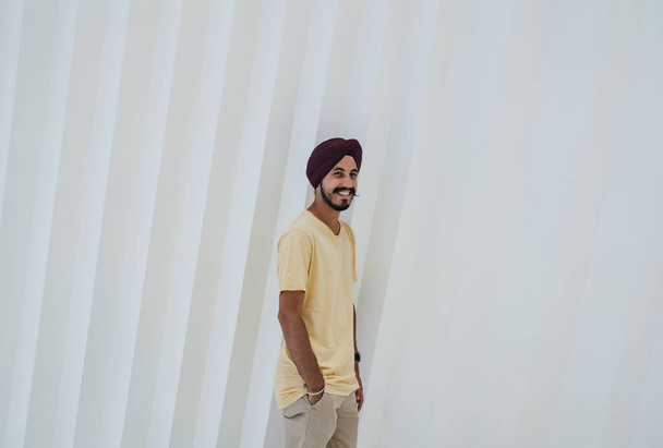 Mosolygó indiai férfi turbánnal pózol a zsebében, miközben a fehér geometrikus fal előtt áll, és a kamerába néz. - Fotó, kép