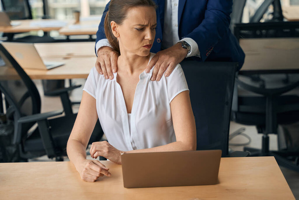 La mujer está usando la computadora portátil en el lugar de trabajo mientras el hombre está haciendo su masaje y coqueteando mientras ella se siente abusada - Foto, imagen