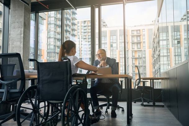 Homem está sentado à mesa com laptop e se comunicando sobre o trabalho com mulher com deficiência - Foto, Imagem