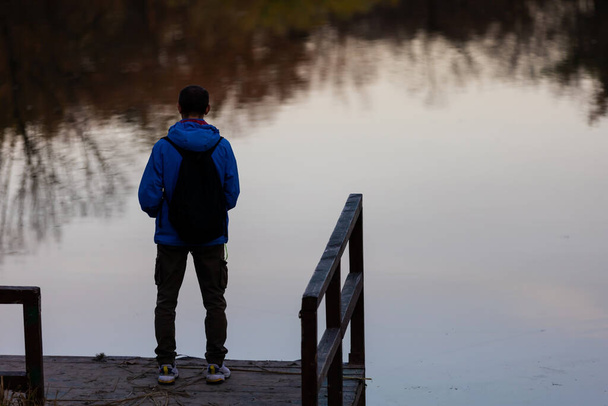 Młody człowiek z plecakiem stojący samotnie na drewnianym kładce i gapiący się na jezioro. Spokojna atmosfera w przyrodzie. Cieszymy się świeżym powietrzem jesienią wieczorem. Widok z tyłu. - Zdjęcie, obraz